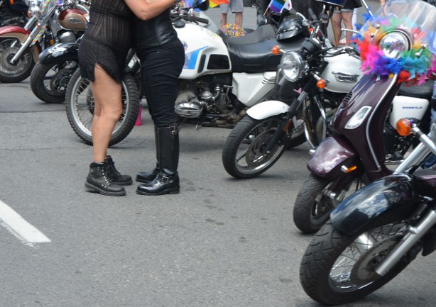 two people hugging, one in black pants and knee high boots, motorcycles parked around them. 
