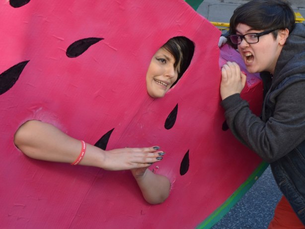 a woman attempts to take a bite out of another woman who is wearing a giant watermelon costume