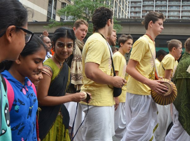 a group of young South Asian women walking in a parade. One of them has her face decorated with paint. In front of them is a group of young men in yellow tops and white bottoms, one has a drum.