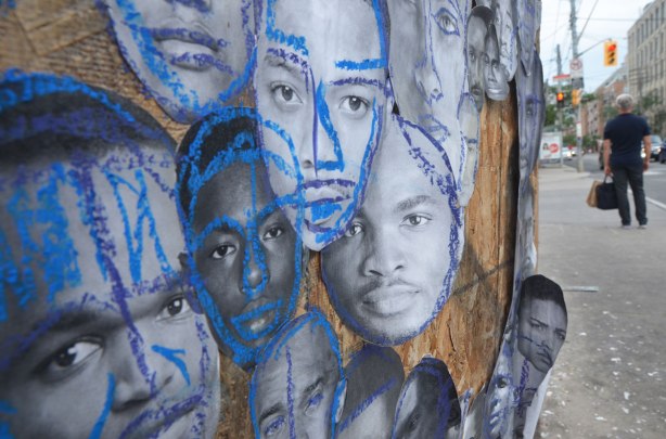 pictures of the faces of black men attached to wood construction hoardings and rough outlines and highlights drawn on the faces with crayon, in red, blue, yellow and green. The faces all look like they are looking into the camera