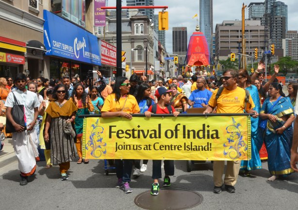 people walk behind a yellow horizontal banner that reads Festival of India, Join us at Centre Island.
