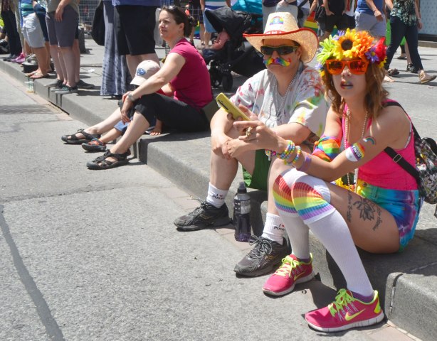 spectators sitting on a sidewalk. A woman with flowers in her hair and pink sunglasses as well as white knee socks. The man beside her has a fedora on. 