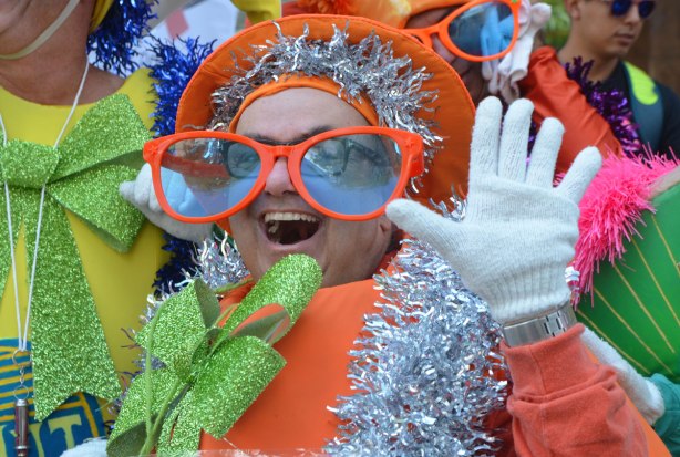 a man dressed as a fruit, an orange, with big green glittery bow in the front and oversized orange frame glasses, also white gloves and an orange hat. He is smiling a big smile and waving at the camera