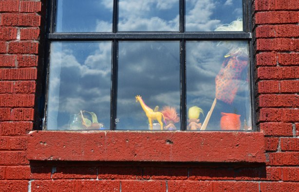 looking into a window, toy yellow giraffe on the window sill as well as two toy trolls, one with yellow hair and the other with orange. Reflections of clouds in the window 