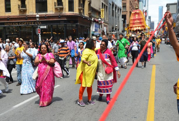 lifting the red rope that separates the parade from the traffic, women dancing and clapping and walking as well as other people, pulling ropes to pull the chariot in the parade
