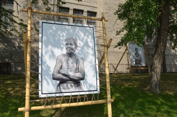 large black and white photo of an older woman with many tattoos, black and white, displayed outside, another portrait in the background