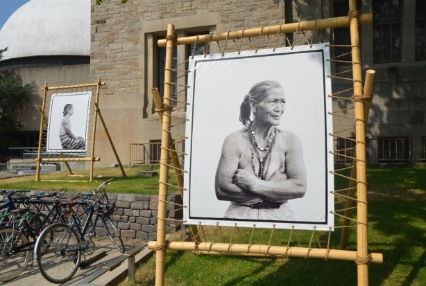 large black and white photo of an older woman with many tattoos, black and white, displayed outside, another portrait in the background