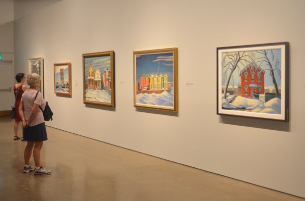 two women look at a line Lawren Harris paintings of brightly coloured houses in winter on a wall in an art gallery