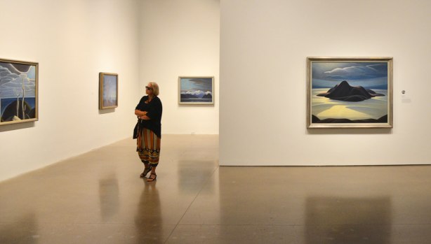 a woman walks through a gallery with paintings on the wall. She stops to look at one of them.