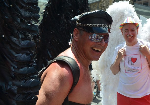 Two men with large feathery wings on their back. The one in front has black wings and is wearing a black leather hat. He is topless. The other man is wearing a white T-shirt and white feathery wings as well as a rainbow striped hat. 