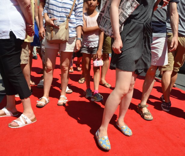 Many legs and feet on a red carpet outdoors. People dressed for hot weather, only face in the picture, a young girl looks bored as she looks at the camera