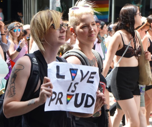 walkers in a dyke march in Toronto - two young women walking together, one is holding a sign that says "love is proud"