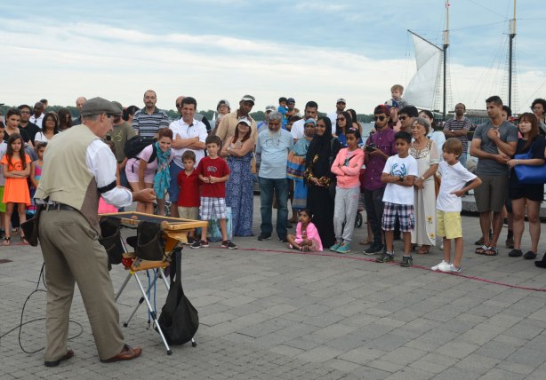 a magician entertains a crowd on the waterfront, many people are watching, men, women and kids