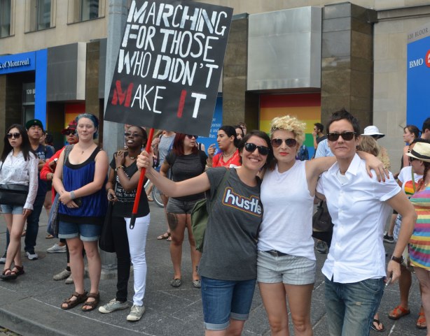 spectators on a sidewalk watching the Dyke March, one woman has a sign that says "Marching for those who didn't make it"
