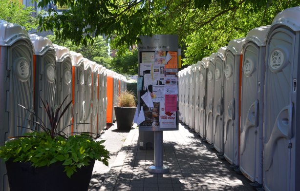 Two lines of Johnny on the spots, facing each other, in the middle is a small message board on a metal pole, outdoors, green leaves above