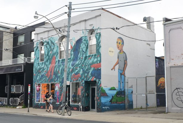 two sides of a building with murals, one side is a large mural of a boy standing on a small island, wearing sunglasses, and saying "Hello", painted by Hebru Brantley and the other side (store front) 