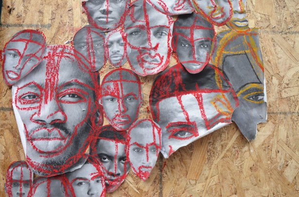 pictures of the faces of black men attached to wood construction hoardings and rough outlines and highlights drawn on the faces with crayon, in red, blue, yellow and green. 