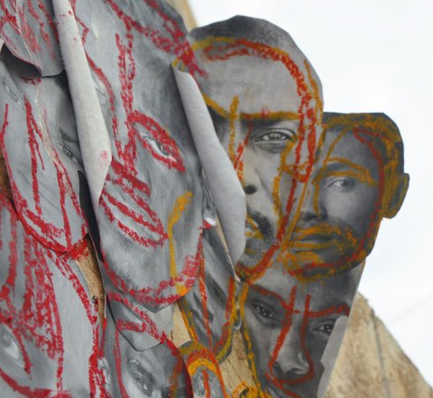 pictures of the faces of black men attached to wood construction hoardings and rough outlines and highlights drawn on the faces with crayon, in red, blue, yellow and green. Some of the faces are peeling away at the edges and the look a bit 3D