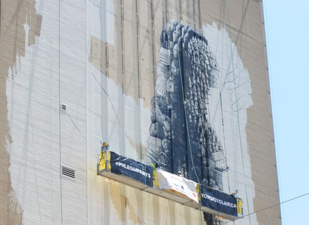 a very large mural on the side of a building is in the process of being painted. An white outline of a seated person with their knees drawn up, one leg has been painted with a design in black