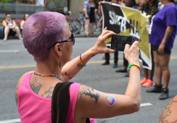 a woman with very short purple hair, a pink sleeveless top, and many tattoos, is taking a picture with her phone of a group marching in the Dyke March 