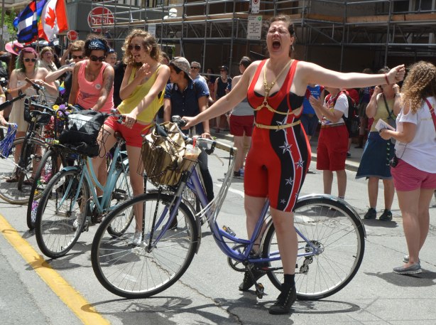 walkers in a dyke march in Toronto - one cyclist in a tight red body suit is yelling in celebration, other cyclists around her