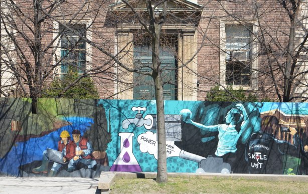 part of a mural on an artwall on hoardings around a construction site for a new engineering building at the University of Toronto, an older building behind, a volleyball player on the mural as well as two students sitting on the ground and looking at a laptop
