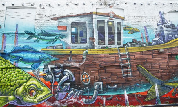 part of a mural by Bruno Smoky across the back of a large building, marine life, lots of fish in blues and greens as well as a small wood boat with a little white cabin on it. 