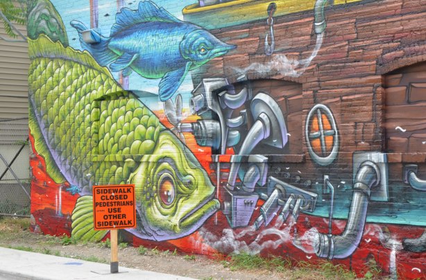 part of a mural by Bruno Smoky across the back of a large building, marine life, lots of fish in blues and greens as well as a small wood boat with a little white cabin on it. 