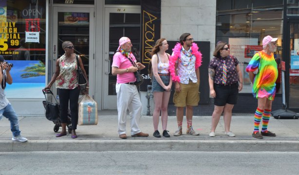spectators on the sidewalk watching a parade. A man with a camera, a woman with her shopping, a man with a pink boa, another man with a pink shirt and white pants 