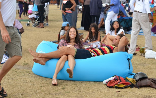 a couple are lounging on a large light blue foamy thing, amongst a crowd at an outdoor music festival