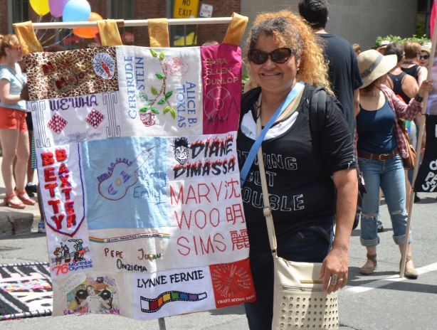 a woman in dark sunglasses stands beside a banner with sections for different famous lesbian women - Mary Woo Sims, Lynne Fernie, Betty Baxter, We Wha, Gertrude Stein, 