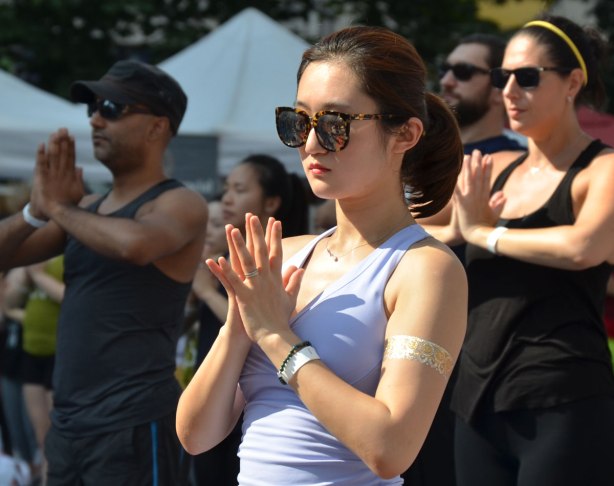 a young Asian woman with big sunglasses, palms together and fingers spread, doing yoga with a group of people outdoors