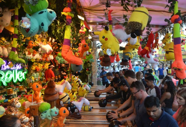 many people playing a midway game at the Ex involving rolling a bowling ball up a short alley. Prizes hand all around them. 