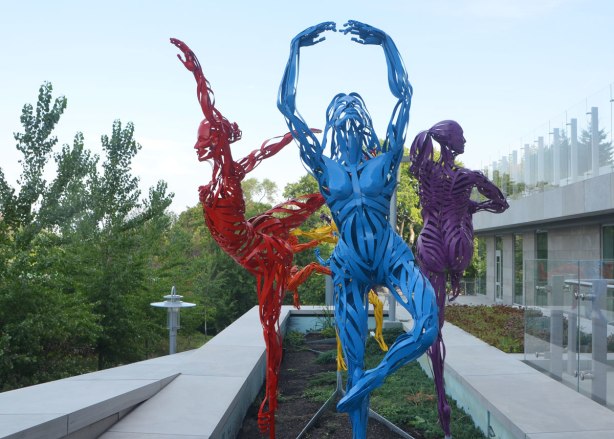 sculptures of three figures in ballet poses, one in blue metal, one in red metal and one in purple. Dancing in a garden outdoors