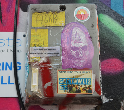 small metal box, part of doorbell system, covered with graffiti stickers, including purple horror face of man