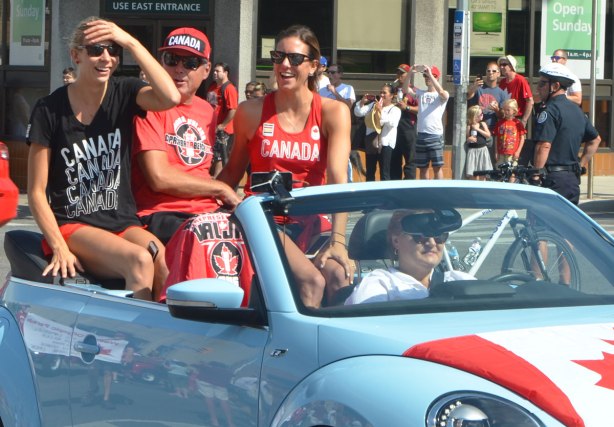 three people sitting in the back of a light blue VW convertible in a parade. They are wearing Canada T-shirts. Man in the middle with a woman on either side. Man is wearing Canada hat. 