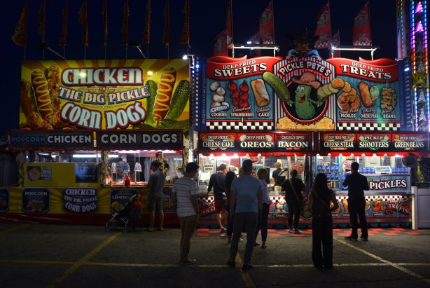 people standing in front of food vendors that ate brightly lit up, selling deep fried food such as deep fried oreos, deep fried pickles, as well as chicken and corn dogs. 
