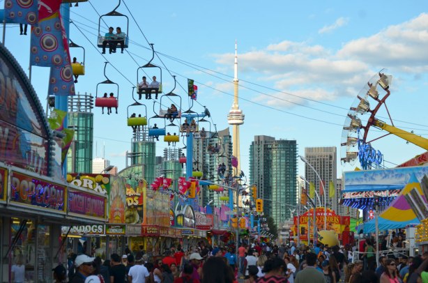 looking down the CNE midway towards the CN tower in the late afternoon as the sun gets low, riders overhead on the Skyline ride, lots of people walking, lots of signs for games and food.
