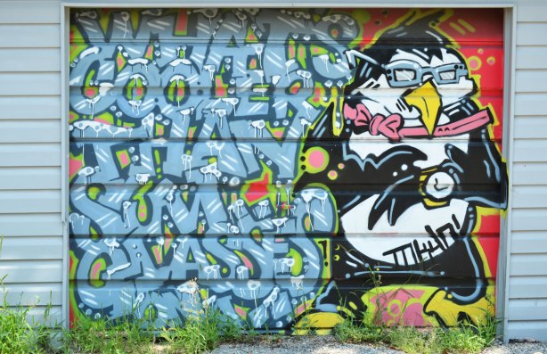 street art mural on a garage door in a lane, a penguin standing upright wearing sunglasses. The words, written in large blue letters with snow on them, What's cooler than sum glasses on?