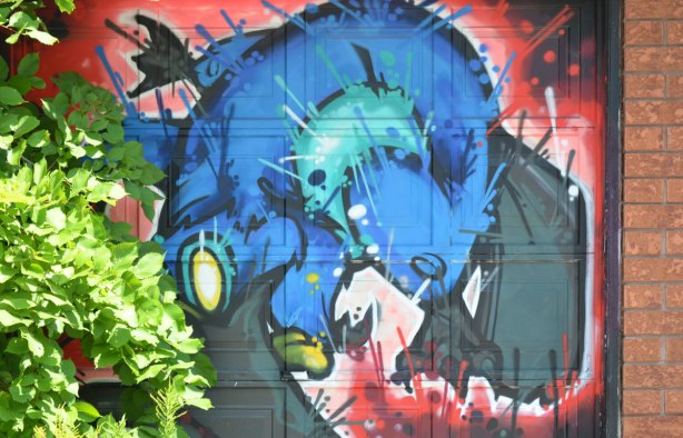 blue animal creature painted on a garage door, green shrub growing in front of part of it, art by cruz 1 in an alley