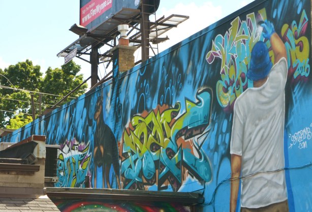 mural on the side of an upper storey, above the roof of the bulding beside it, a dag, some text, and the back of a man painting on the wall 