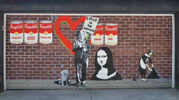 copies of famous street art painted on a garage door. Brick background has been painted on too. Mona Lisa, Campbell soup cans, the woman lifting the corner of the wall to sweep things under it. Einstein holding a placard that says love is the answer. A little dog by Albert Einstein's feet. 