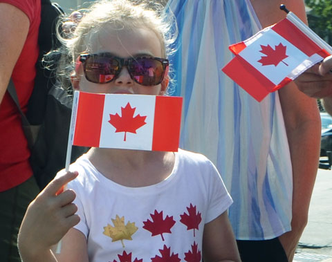 a young girl in a t shirt with yellow and red maple leafs on it, also wearing large sunglasses, holds a small Canadian flag in front of her mouth 