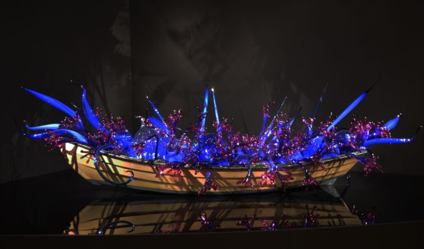 a wooden boat on a black reflective surface. The boat is filled with blue and magenta pieces of sculpted glass