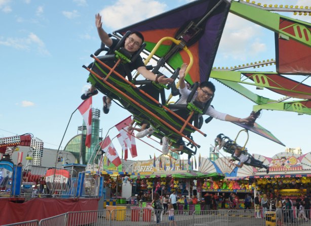 Two young Asian men on a ride, they are lying on their stomachs and appear to be flying, in circles, with the ex midway behind them