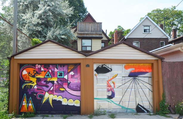 two single car garages in an alley, both with street art paintings on them, also the back of the houses behind them in the lane. 