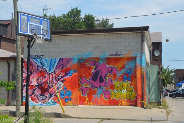a low concrete building in an alley with street art by Cruz 1 art, one says girl power. Large pink skull, basketball hoop in the foreground. 