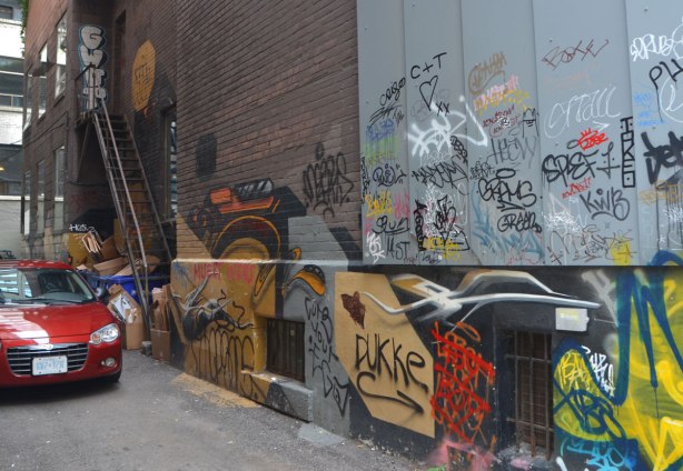 a red car is backed into an alley, and is parked there, along the wall beside the car is a lot of graffiti spray painted and written on the wall. There is a stair case in the back part of the wall.