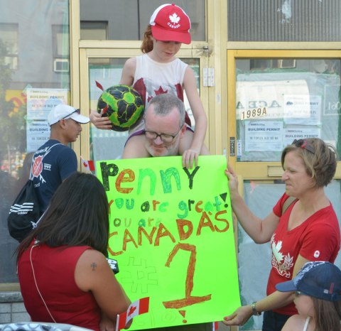 A girl sits on her father's shoulders as they show a sign that they've made to other people who have come to watch a parade. The sign says Penny you are the greatest. Canada's number 1. 