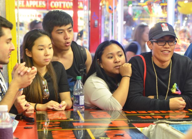 a group of young people around a counter playing a game of chance outdoors, at a fair, the cne